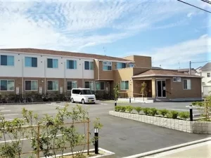 介護施設の外観。駐車場と白い車、玄関エントランスが見える