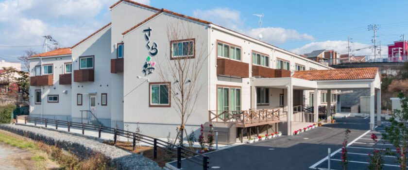 白とレンガ色の外観が特徴的な介護施設の建物全景。広い敷地に立つモダンな構造。