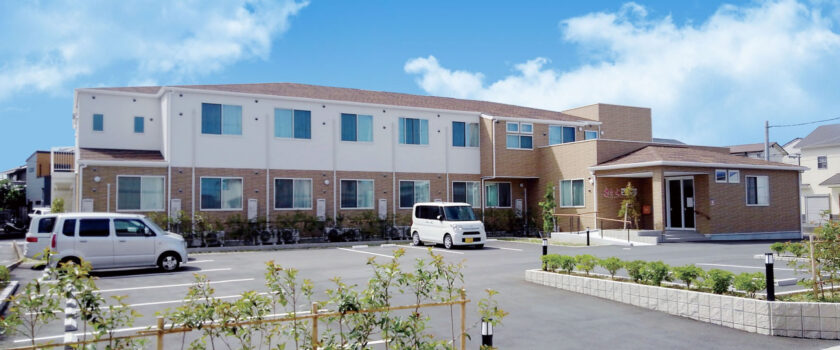 介護施設の建物外観。白と茶色のツートンカラーの2階建て施設。駐車場に複数台の車が停まっている。
